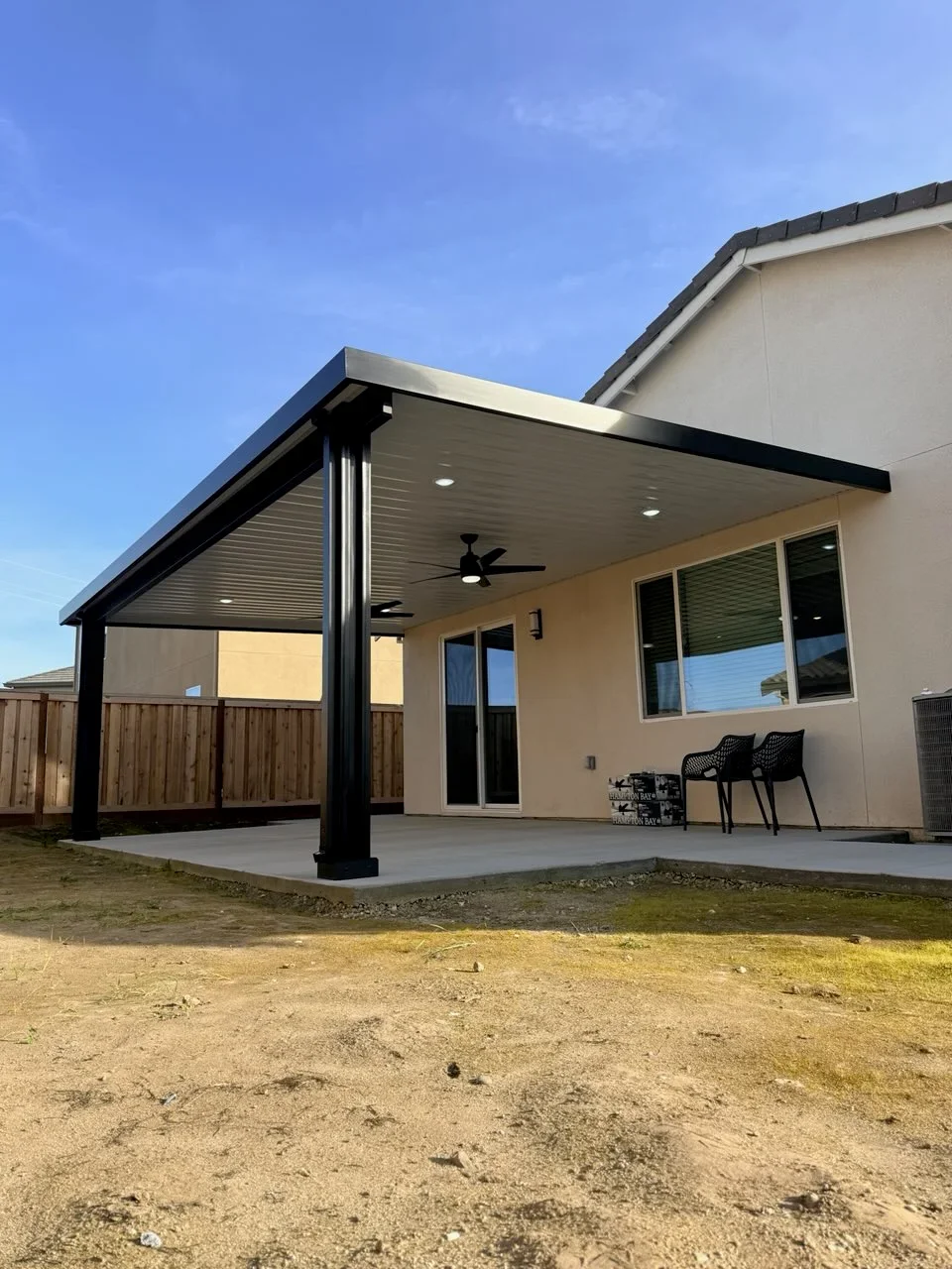 Black aluminum louvered gazebo with recessed lights, ceiling fan, and wooden privacy screen attached to house