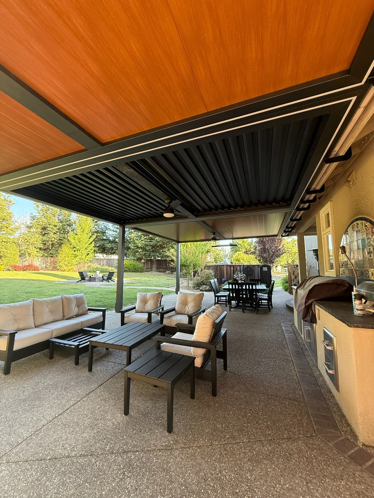 Black louvered gazebo with aluminum frame over concrete patio, seating area with cushioned furniture, outdoor kitchen visible