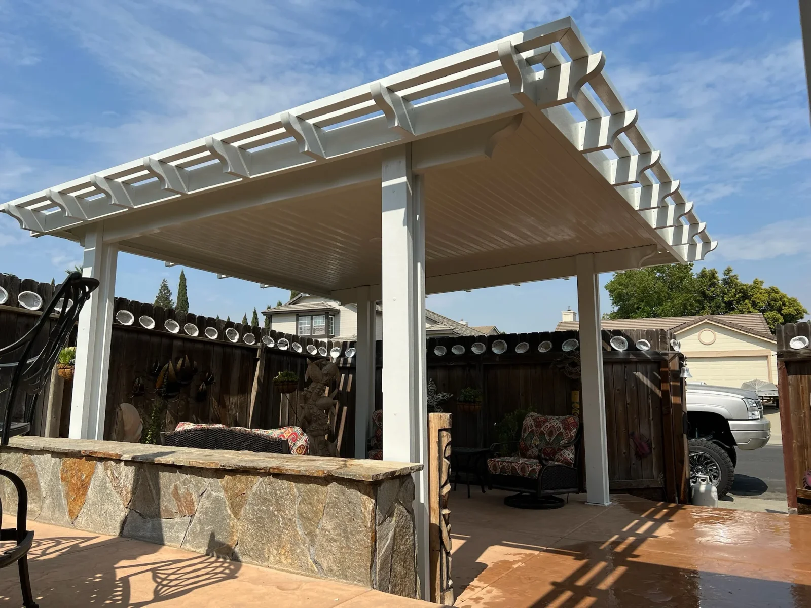 White aluminum pergola with adjustable louvered roof over patio with stone base and dark wood fencing